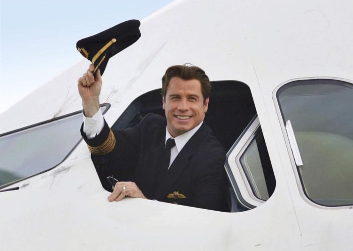 John Travolta smiling and waiving his cap from an open window in an airplane's cockpit, Photo Credit: johntravolta/Instagram